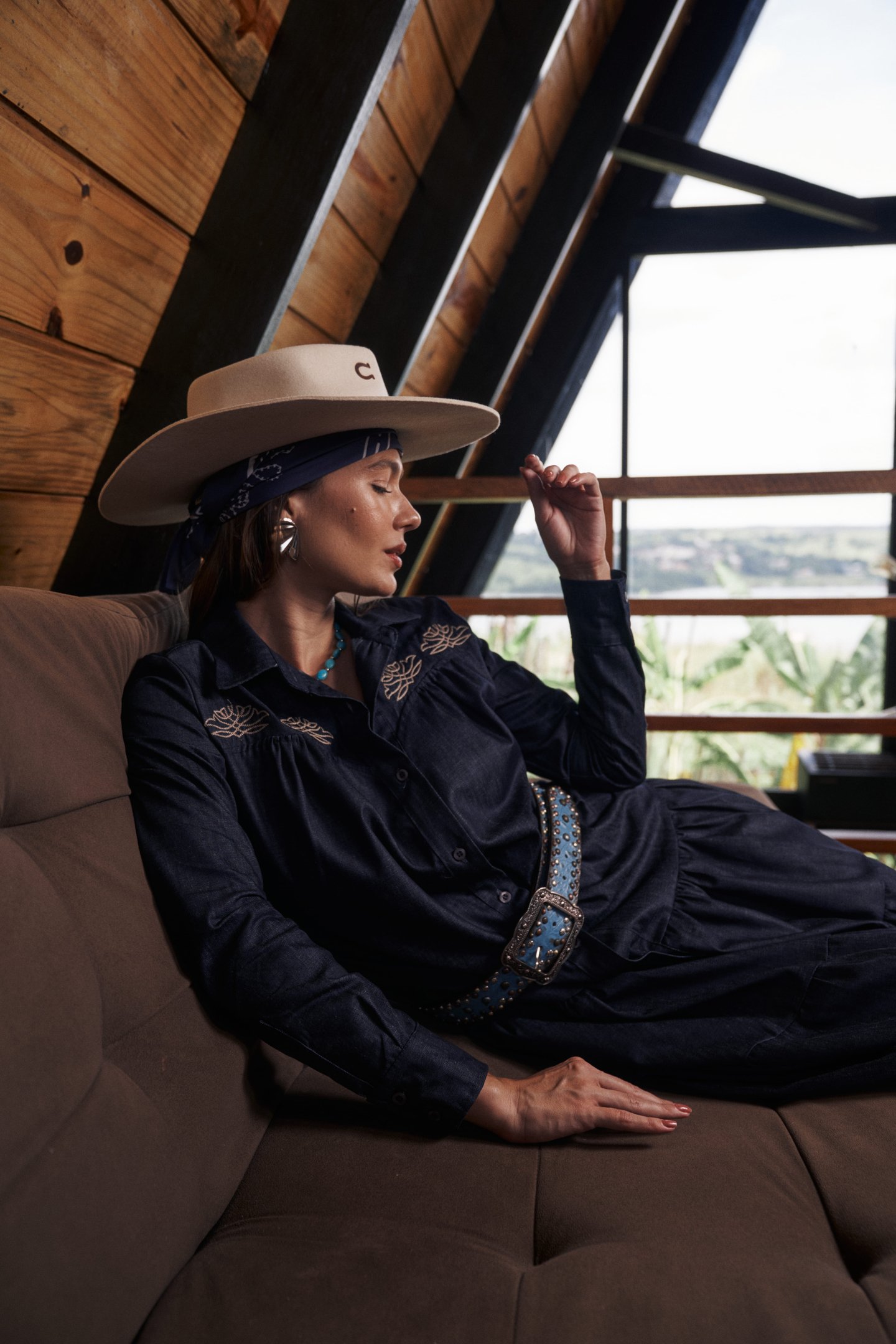 Pessoa relaxando em um sofá, usando chapéu e roupa estilo country, com janela ao fundo.