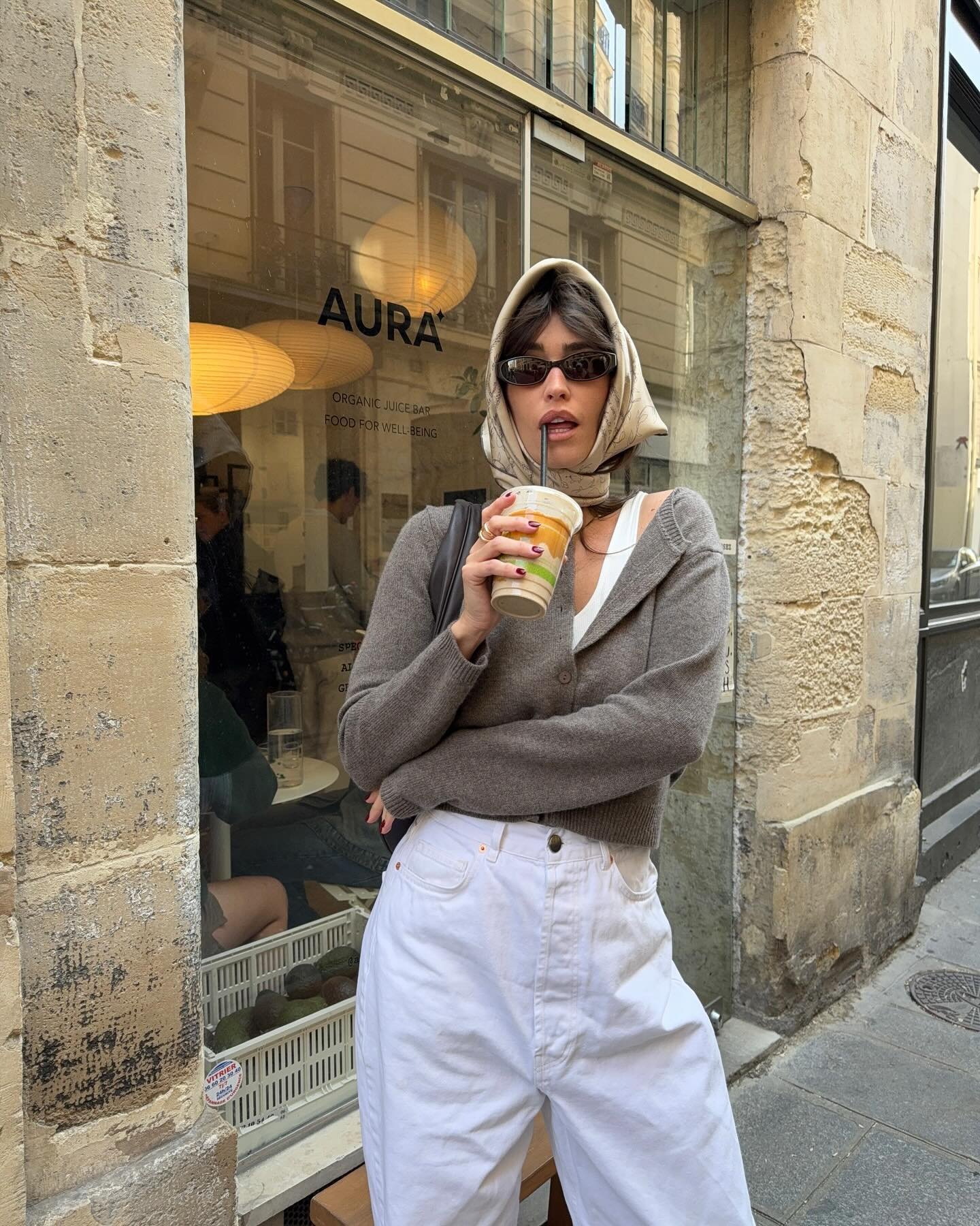 Pessoa com look casual de moda outono inverno 2025, usando óculos escuros, lenço na cabeça e segurando bebida.