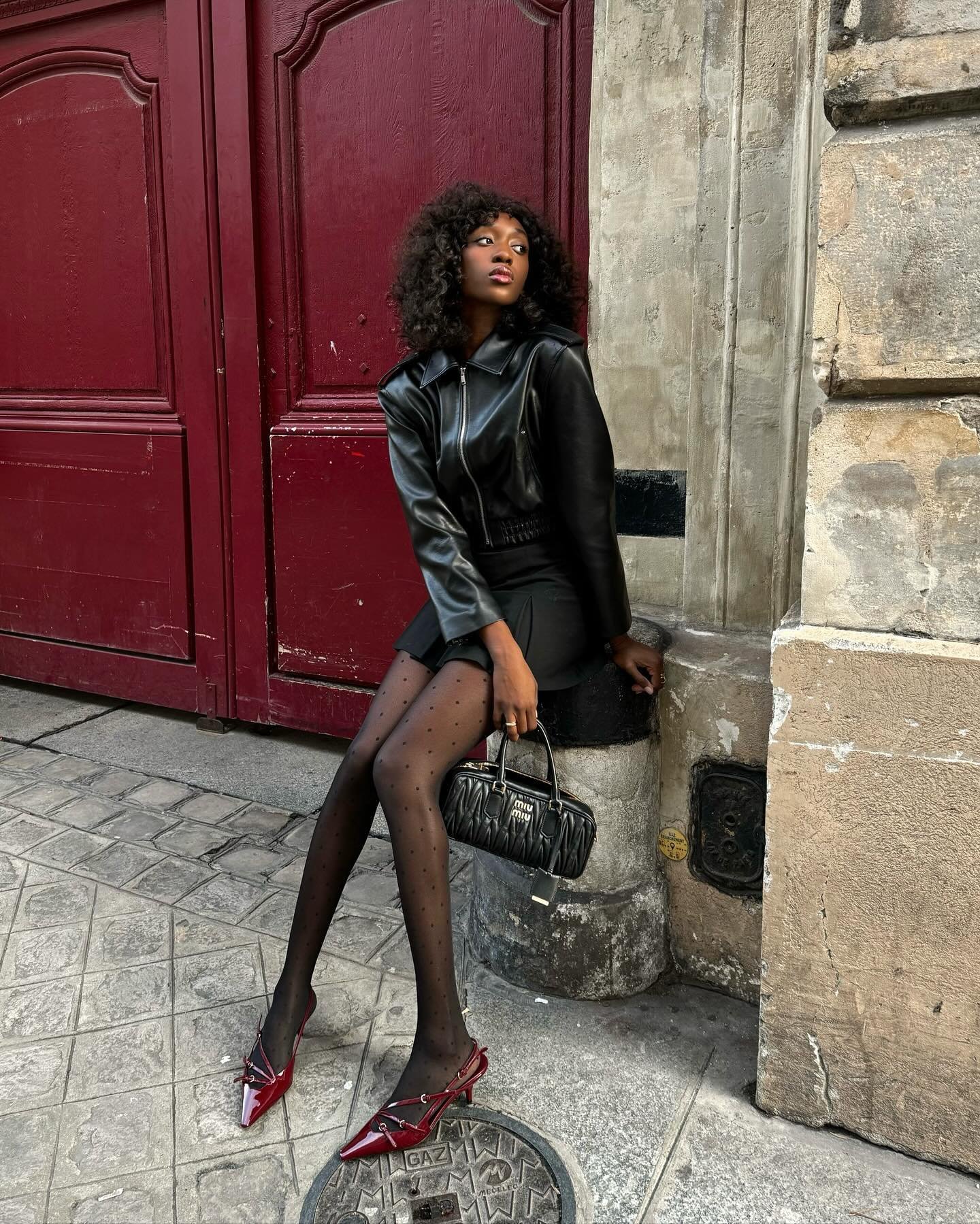 Jovem com jaqueta de couro, meia-calça preta e sapatos vermelhos, em frente a portão vermelho. Bolsas e moda outono inverno 2025.