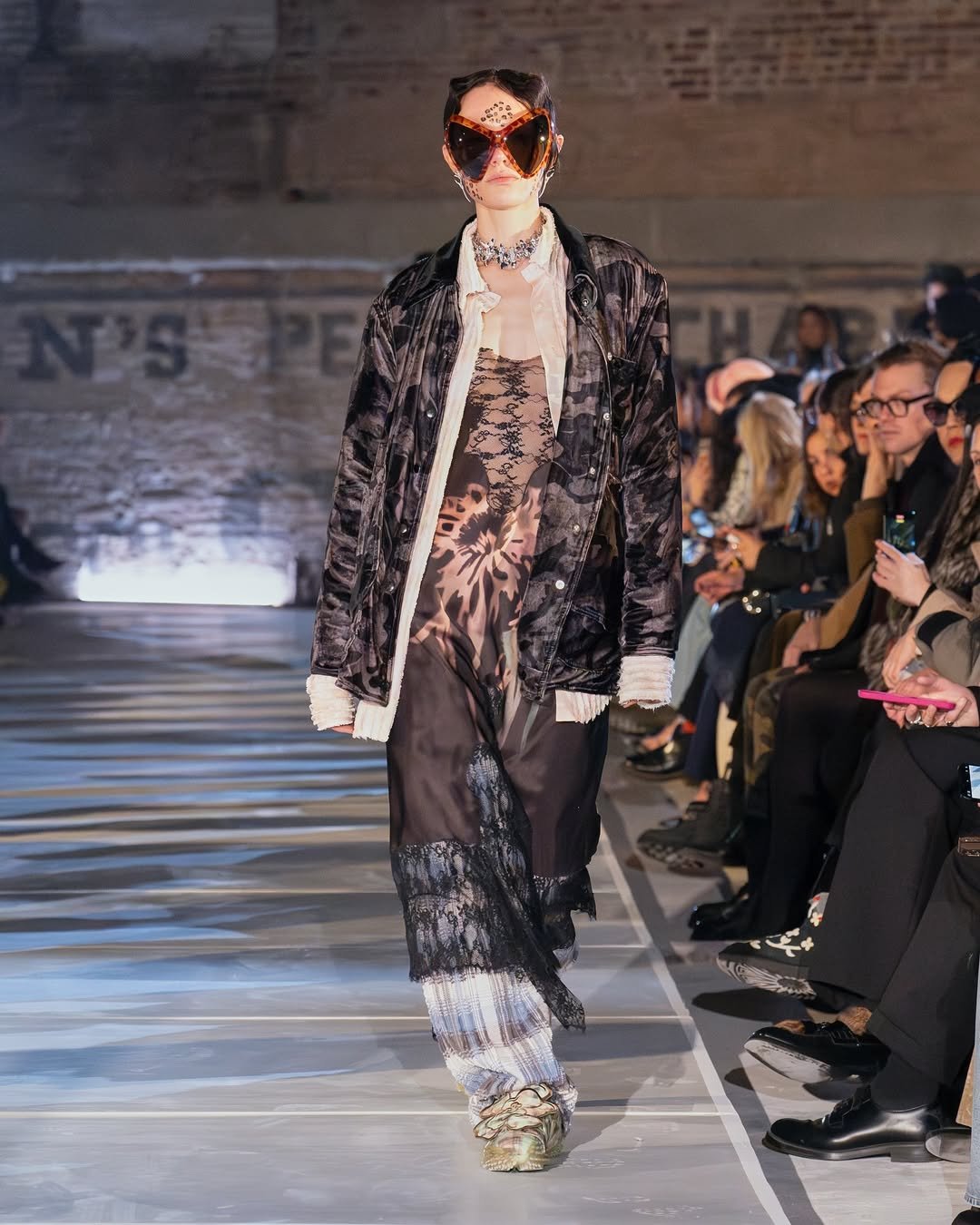 Modelo em desfile, vestindo look com sobreposições e estética inspirada na moda Y2K, incluindo óculos chamativos.