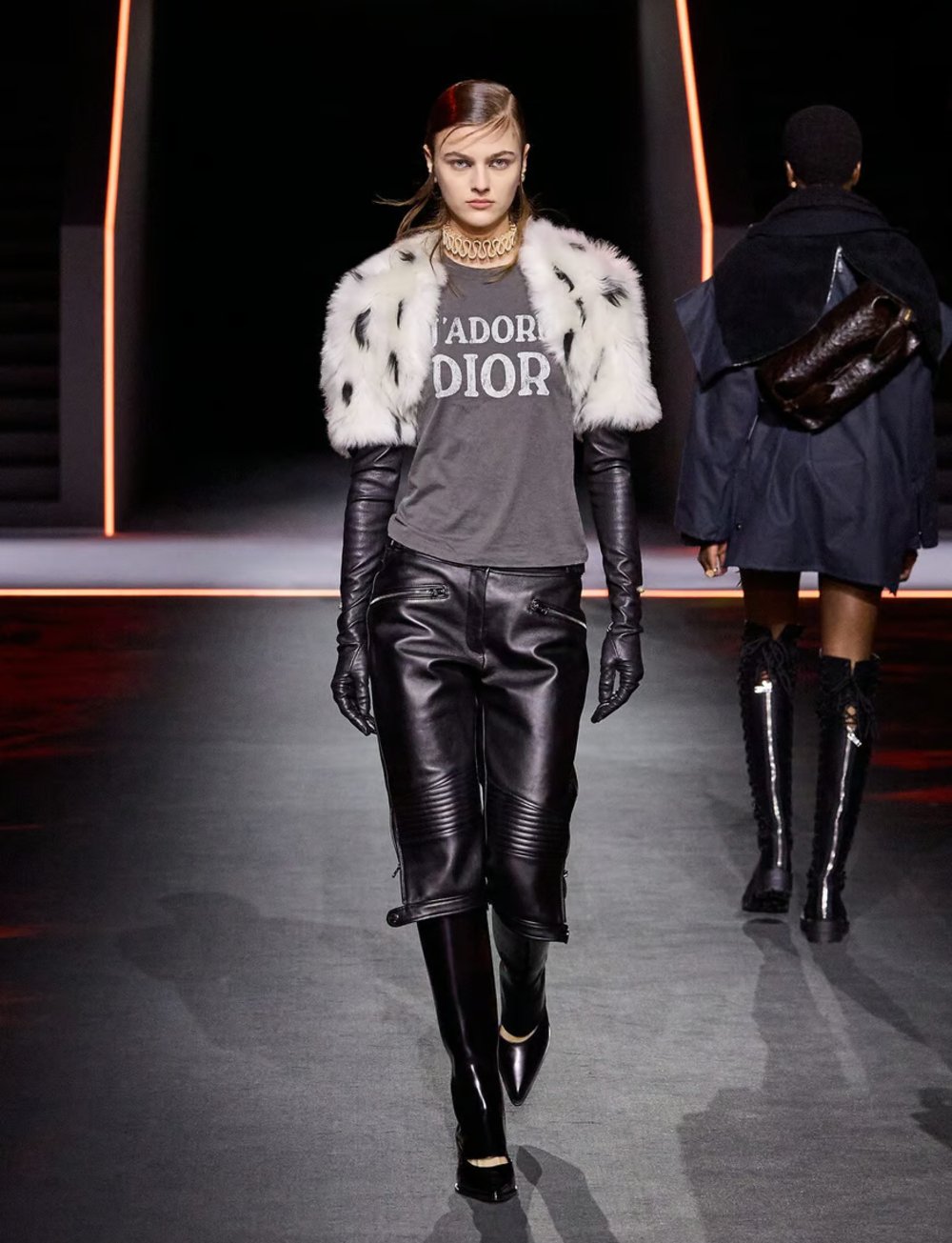 Modelo em passarela com look inspirado na moda Y2K, usando casaco de pele sintética, camiseta grafite e calça de couro preta.