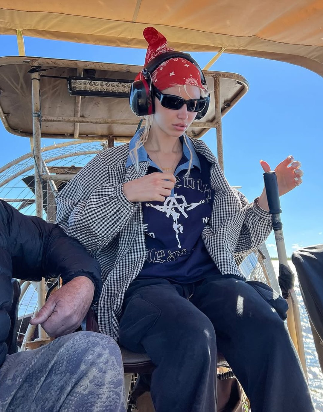 Pessoa sentada com bandana vermelha e fones de ouvido, vestindo óculos escuros e camisa estilo moda Y2K xadrez.