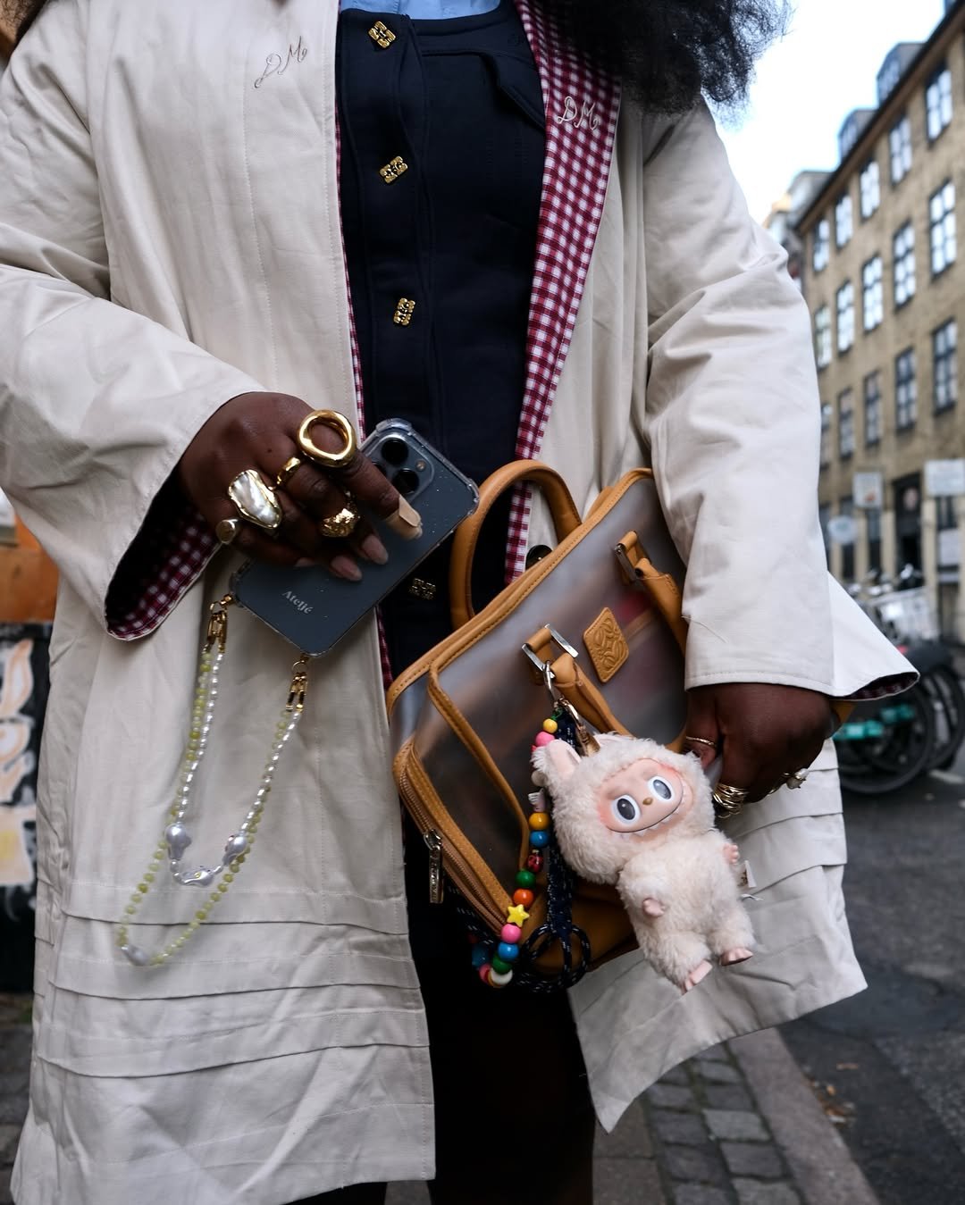 Casaco de padrão xadrez interno, bolsa com chaveiro de pelúcia e anéis chamativos em um estilo eclético.