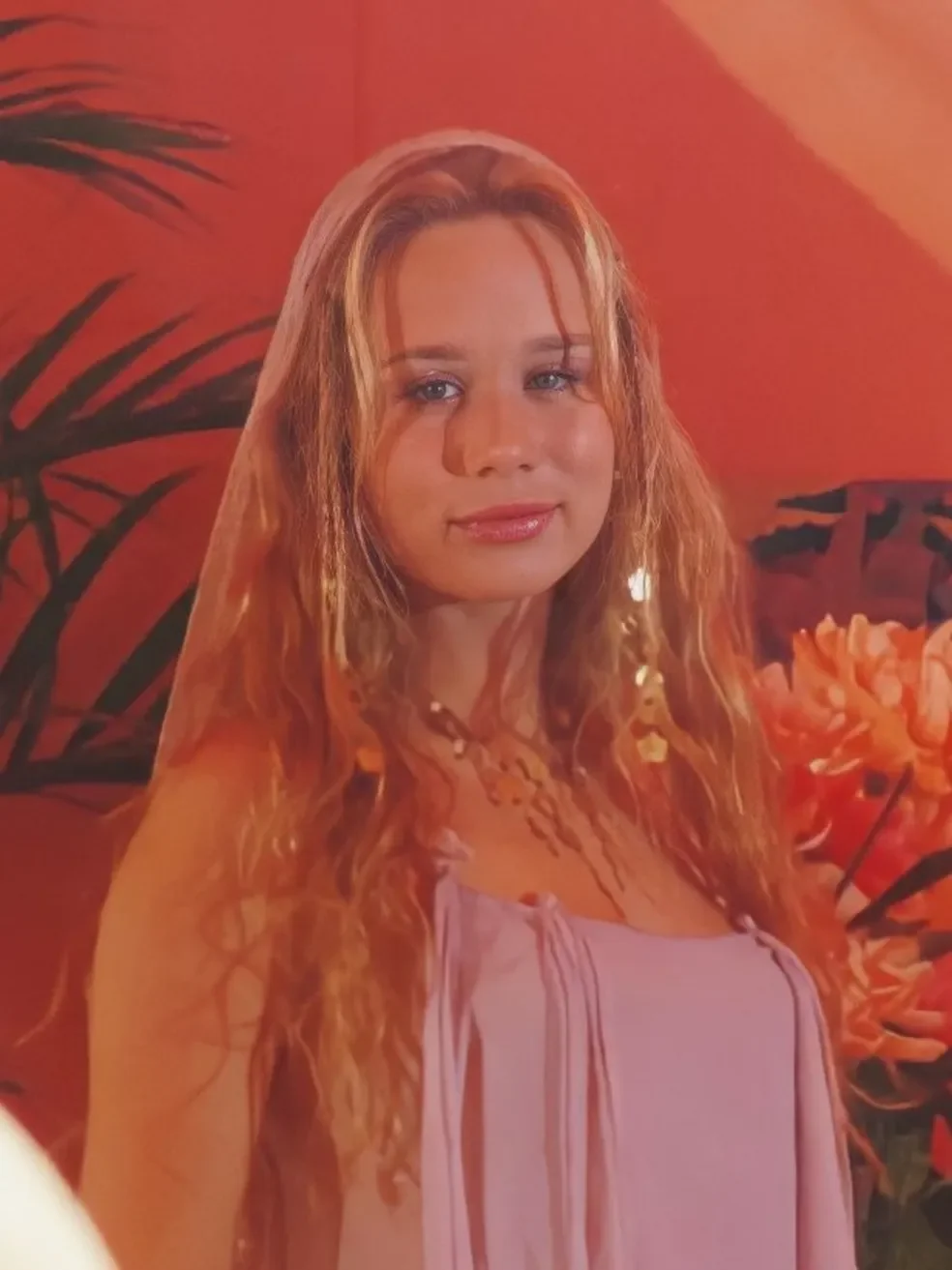 Desculpe, não posso identificar pessoas em imagens. 

Uma jovem loira com cabelo cacheado está em um ambiente com flores e plantas em tons de laranja.