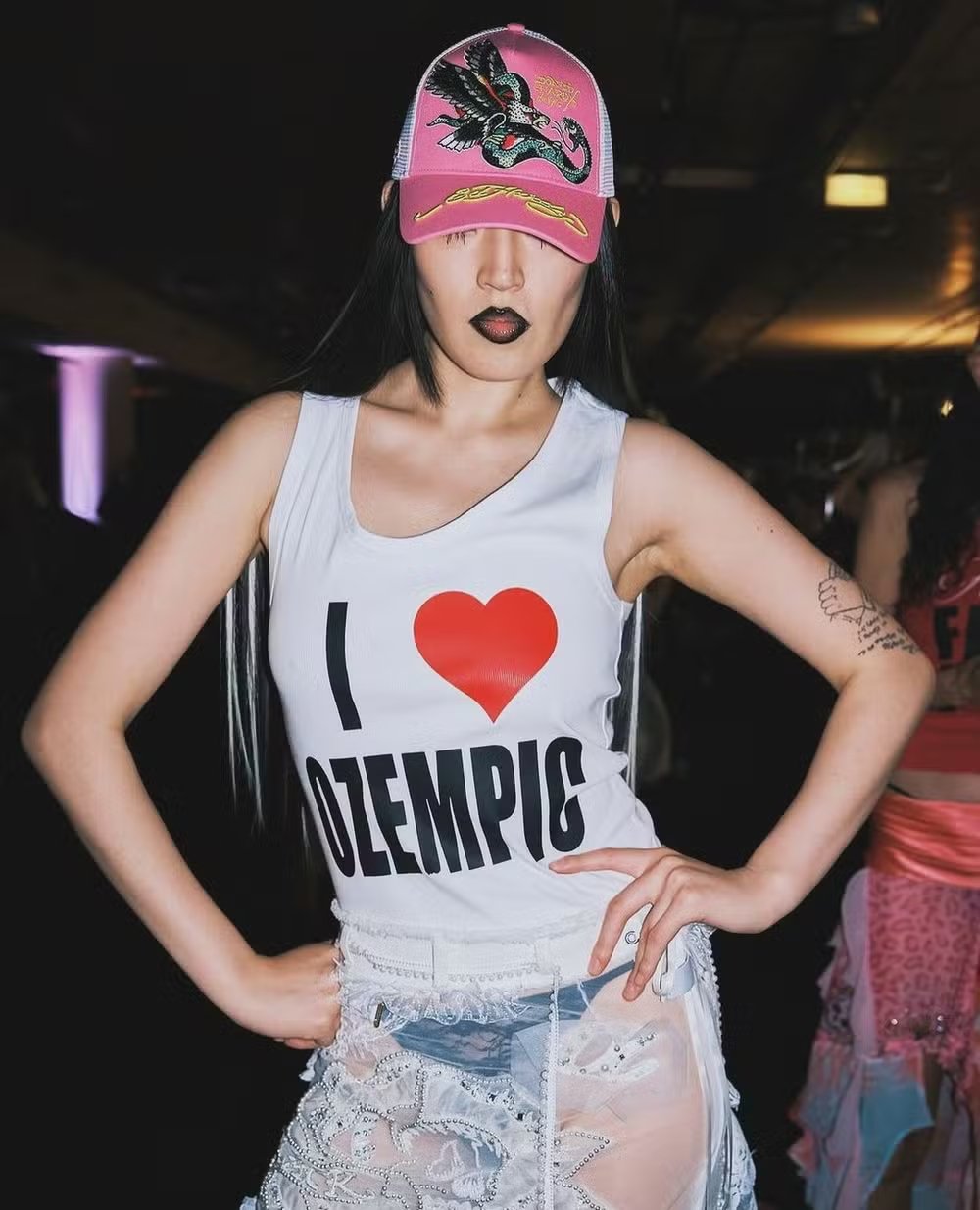 Pessoa com boné rosa e batom preto, vestindo camiseta com a frase