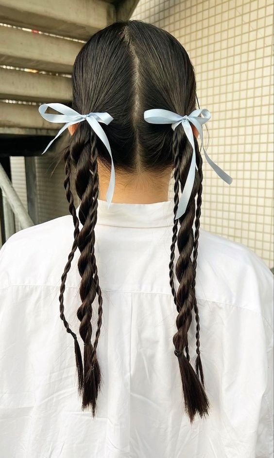 Tranças com laços azuis decoram cabelo escuro dividido ao meio, criando um elegante estilo simétrico.
