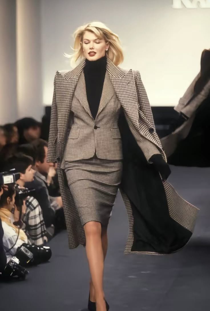 Modelo desfilando com um terninho xadrez, composto por saia e casaco combinando, em uma passarela de moda.