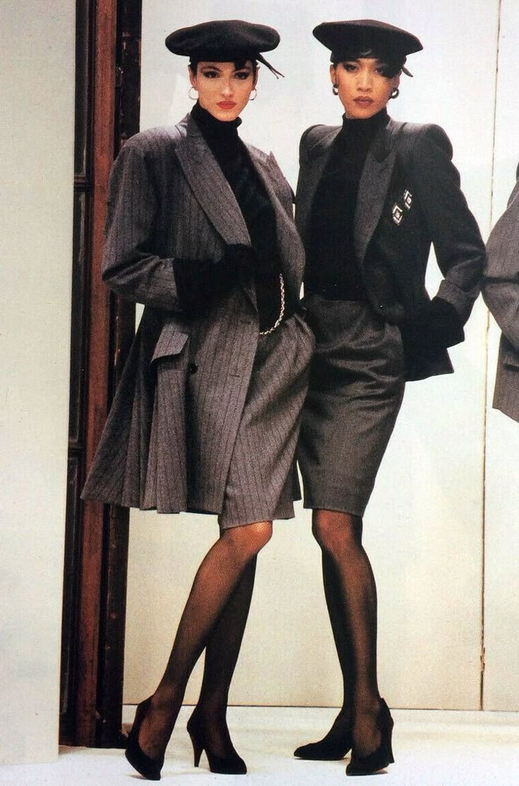 Duas mulheres em terninhos cinza com boinas, usando botas de salto, posam lado a lado.