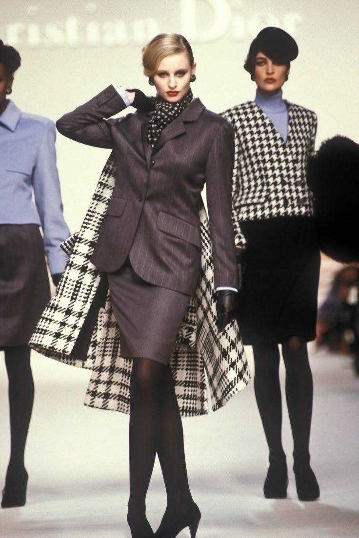 Modelo desfilando com terninho cinza e casaco quadriculado, acompanhada por outras modelos com trajes elegantes.