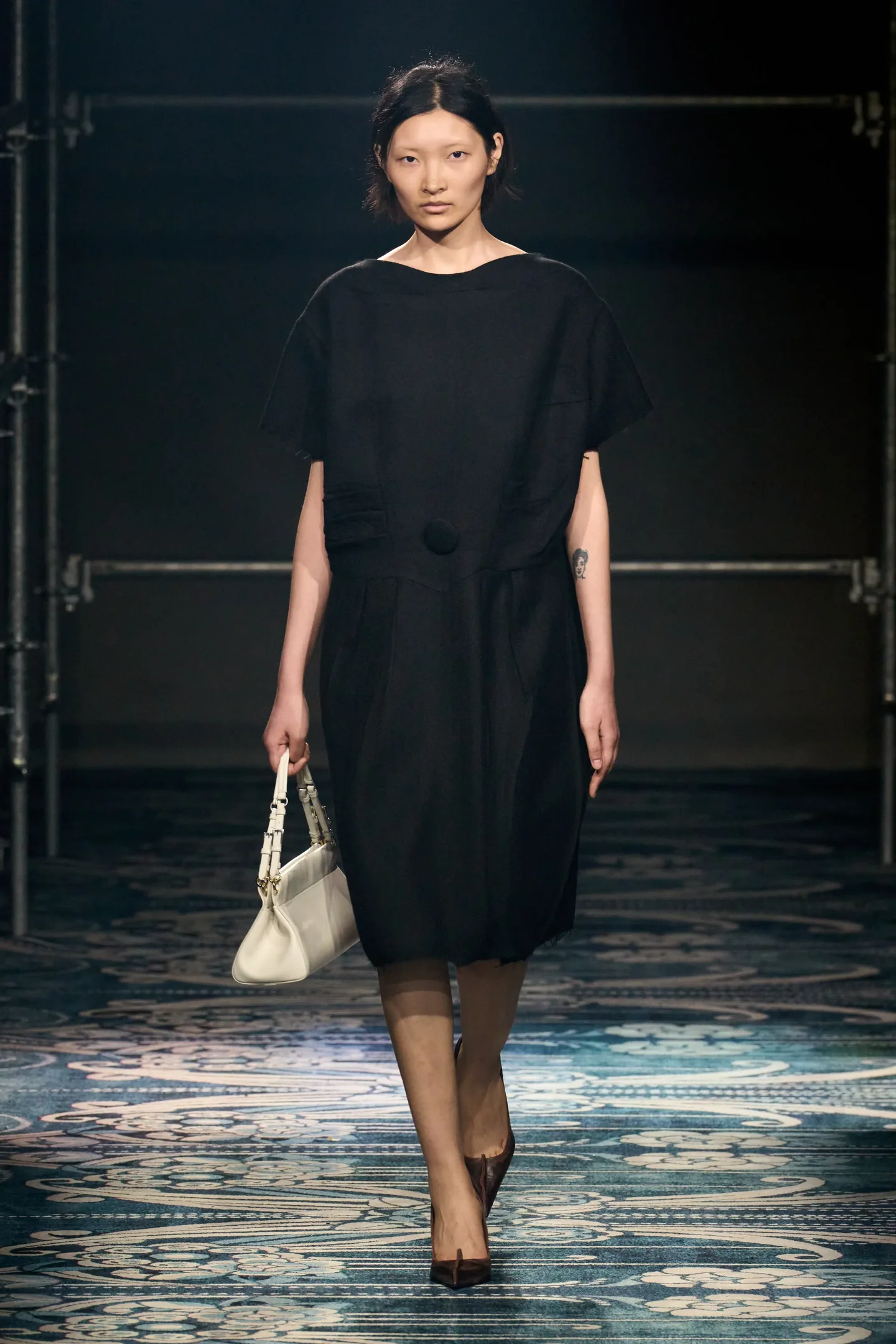 Modelo com vestido preto elegante e bolsa branca, exibe visual conservador na passarela.