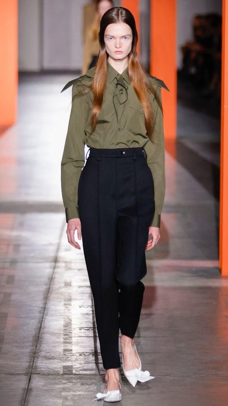 Modelo desfilando em calça preta elegante e camisa verde militar, com sapatos brancos, em passarela moderna.