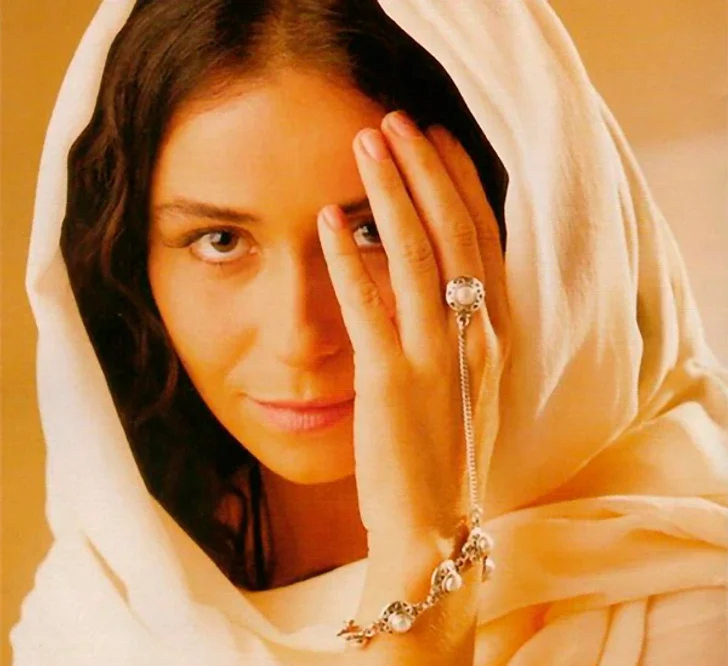 Mulher de lenço bege cobrindo a cabeça, com mão no rosto e pulseira destacada. Estilo lembra personagem de novelas brasileiras.
