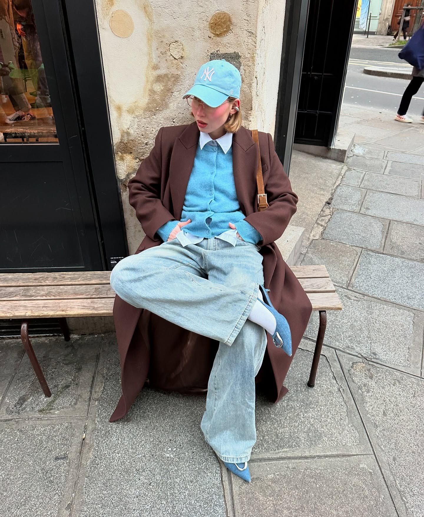 Pessoa usando roupa de frio: casaco marrom, suéter azul, jeans largos e boné azul, sentada em um banco.
