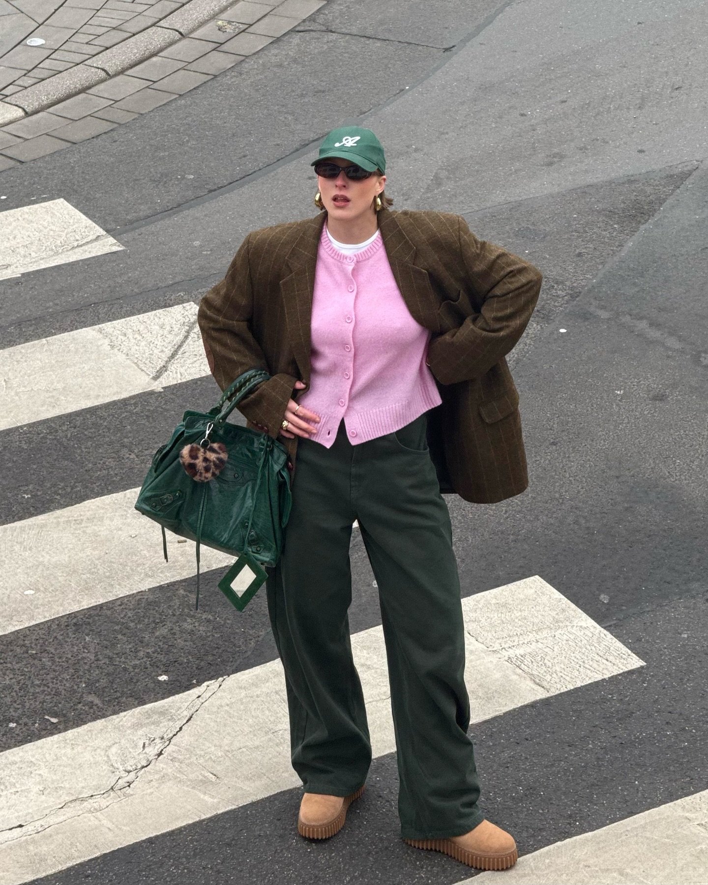 Pessoa em roupa de frio, com casaco marrom, suéter rosa, calça verde e boina, em uma faixa de pedestres.