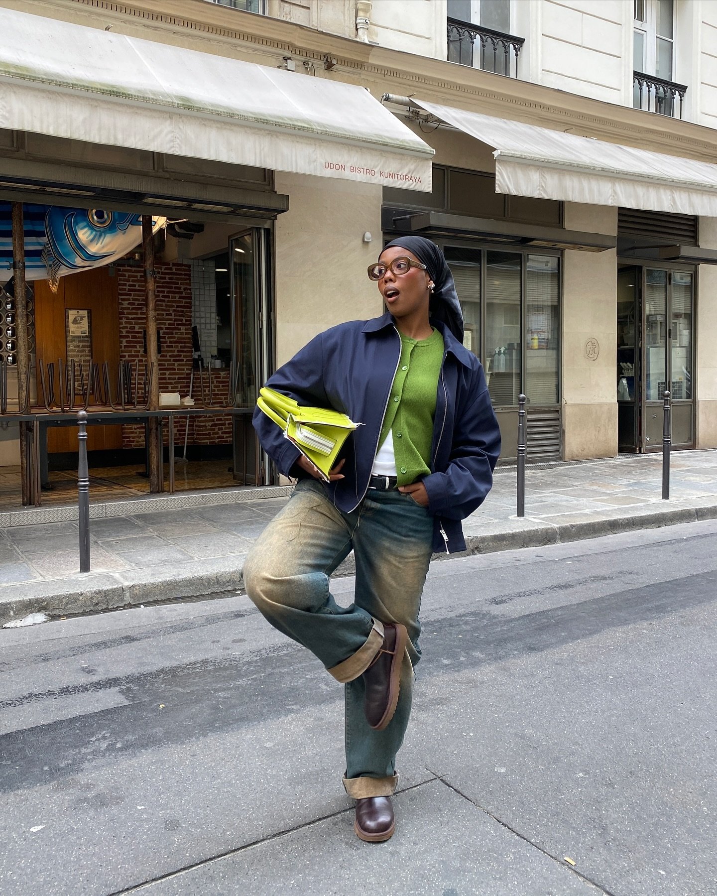 Pessoa usando roupas de frio estilosas, incluindo jaqueta azul escura e cardigã verde, segura uma pasta verde em pé na rua.