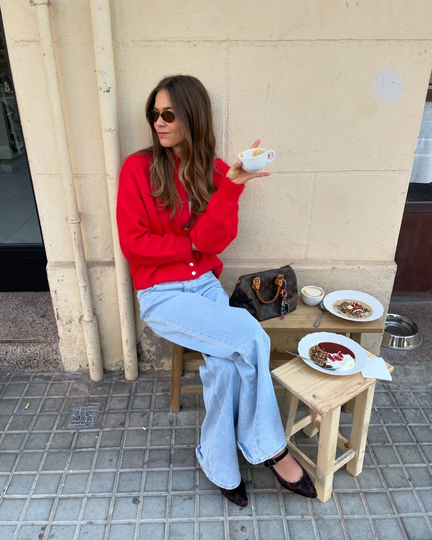 Mulher com suéter vermelho, peça de roupa de frio, segura xícara sentada em ambiente externo perto de pratos de comida.