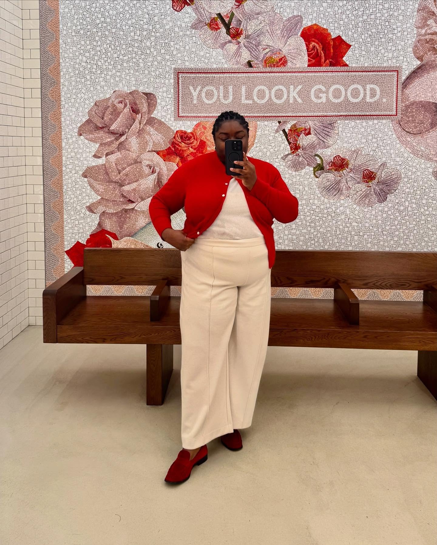 Pessoa vestindo roupa de frio com cardigan vermelho e calça bege. Fundo florido com a frase 