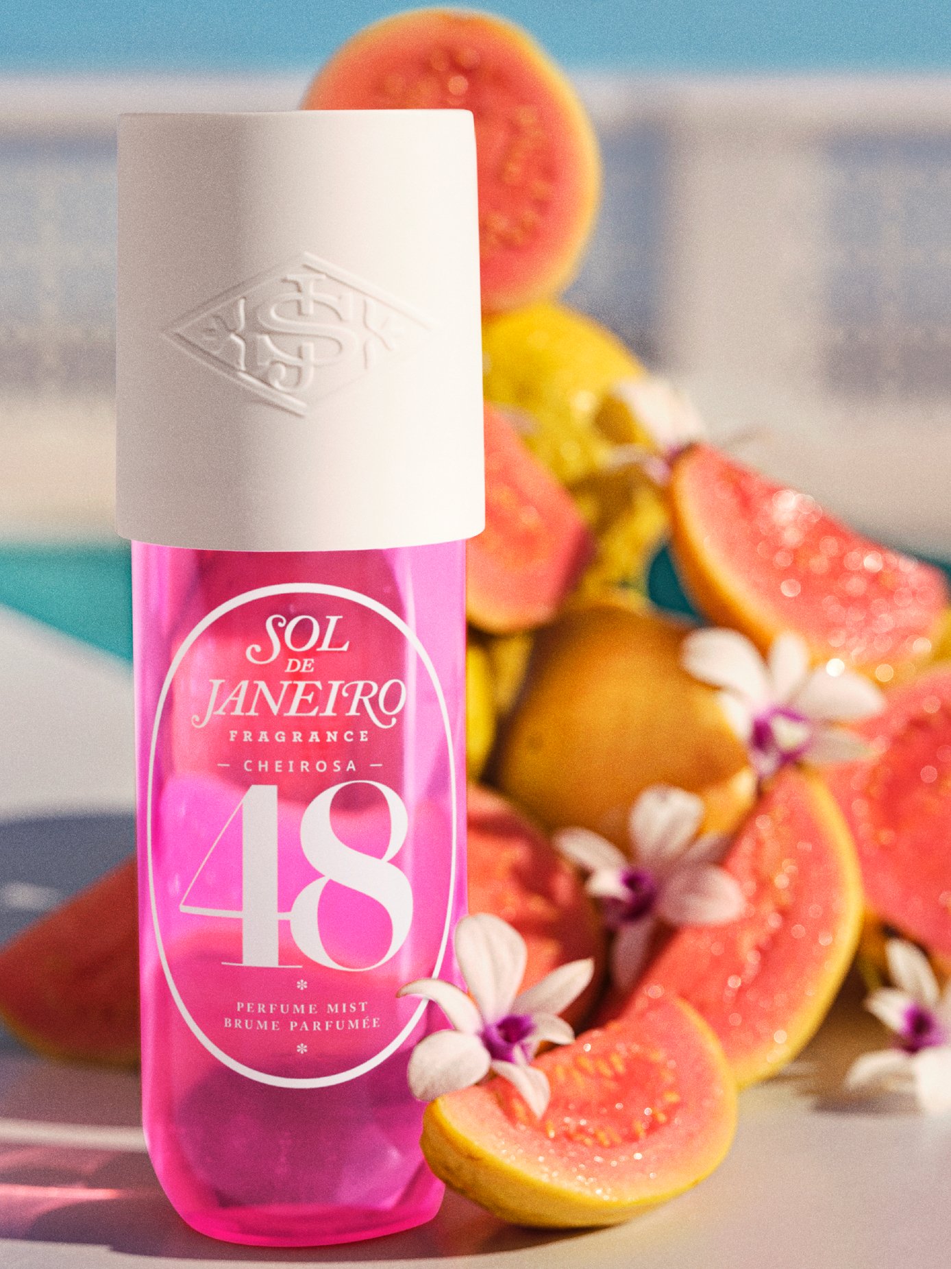 Perfume mist rosa Sol de Janeiro, cercado por frutas tropicais e flores delicadas. Lançamentos de beleza de abril.