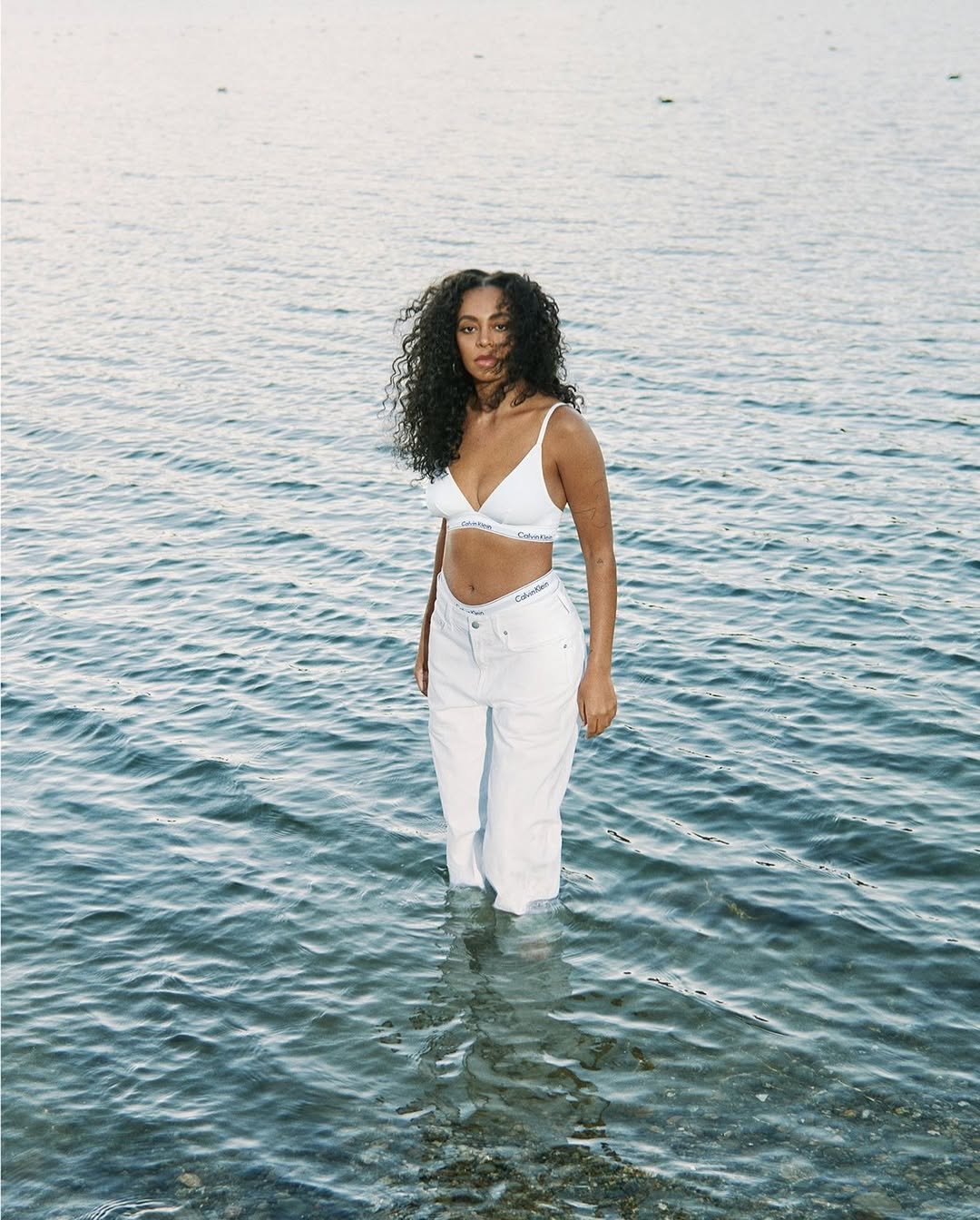 Jovem em pé na água, vestindo roupas brancas, cenário de lago ao fundo. Solange Knowles