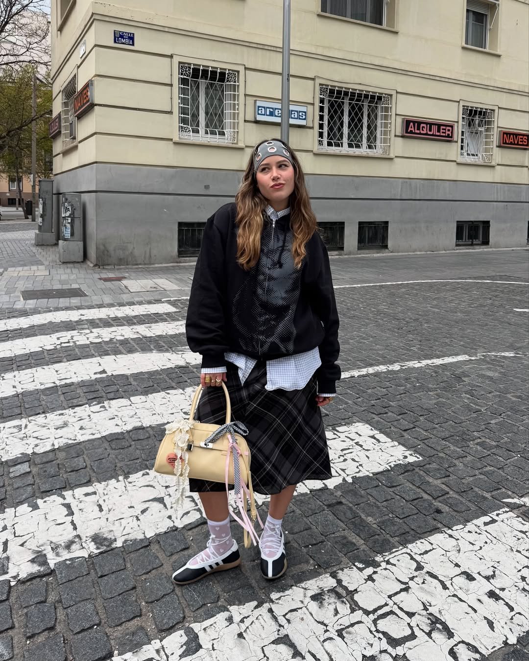 Jovem com saia xadrez e bandana, segura bolsa clara e calça tênis bailarina, parada em faixa de pedestres.