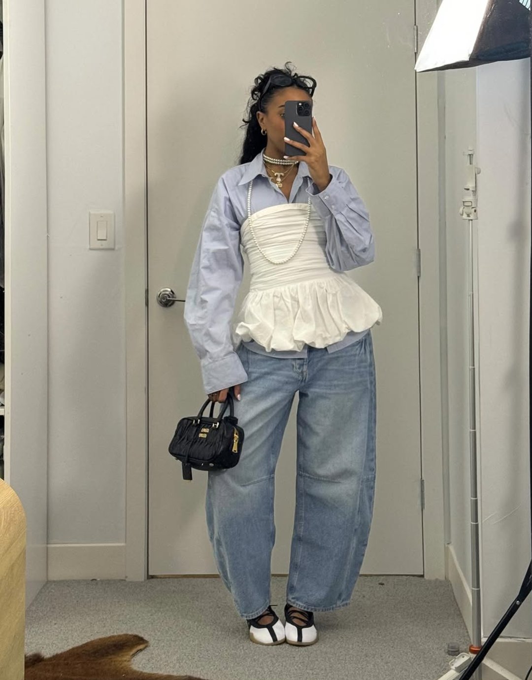 Pessoa com camisa azul, jeans largos e corpete branco posando em frente ao espelho. Segura bolsa preta e usa calçados estilo tênis bailarina.