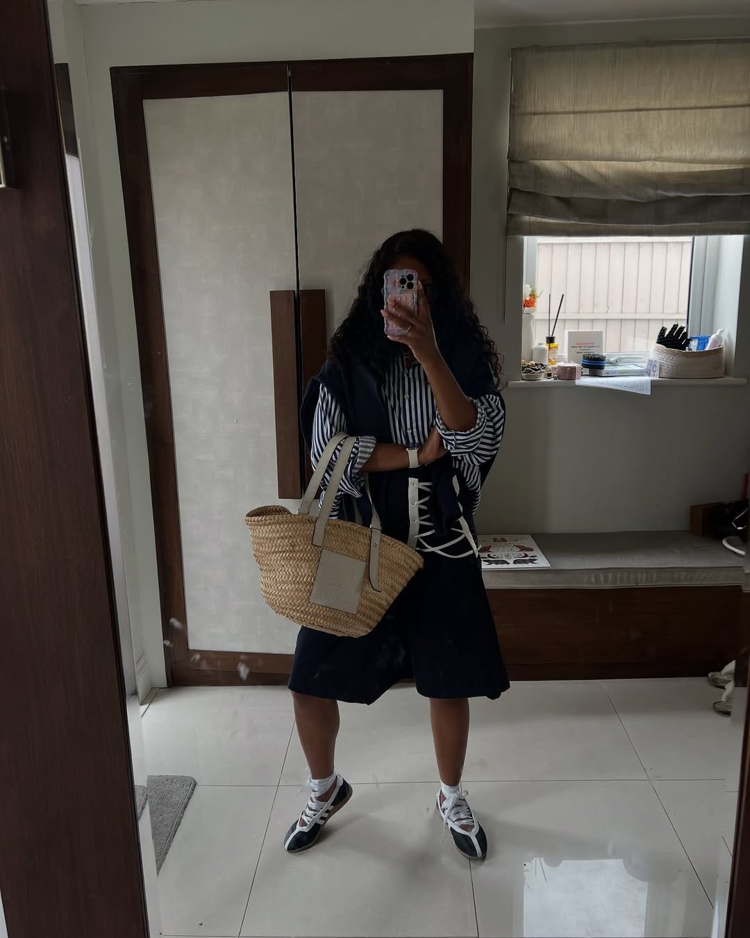 Pessoa com camisa listrada, saia escura e tênis inspirados em sapatos de bailarina, segurando bolsa de palha.