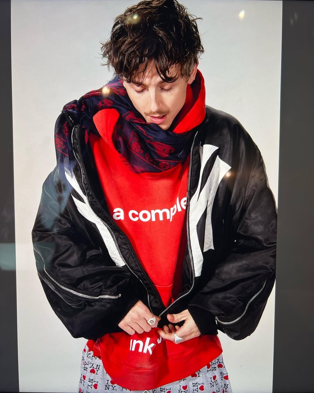 Pessoa veste moletom vermelho, jaqueta preta oversized e lenço. Estilo lembra elementos da moda Y2K.
