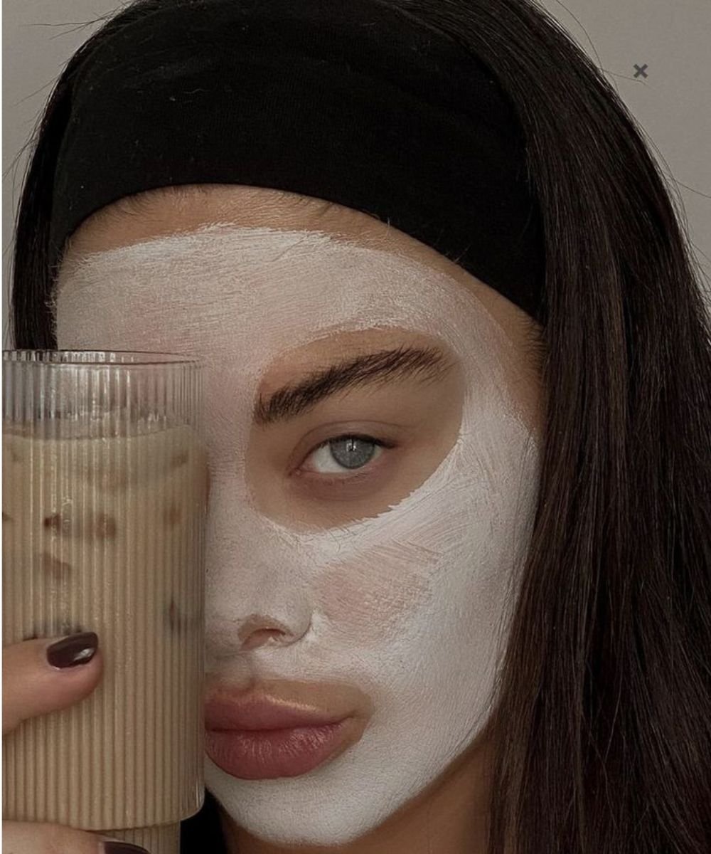 Pessoa com máscara de argila branca no rosto, usando roupão, olhos fechados e cabelo preso.
