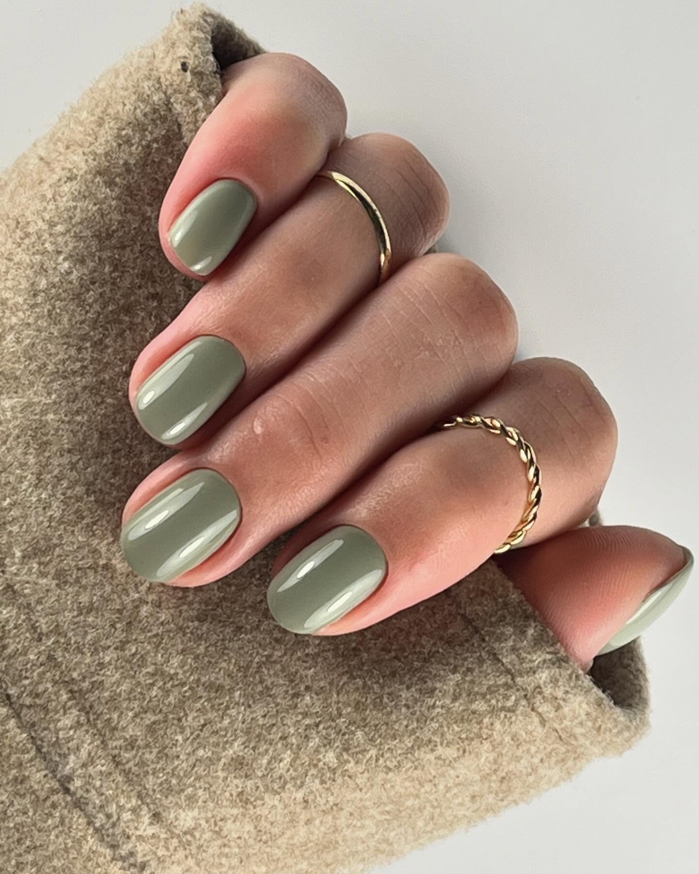 Unhas com esmalte verde oliva e anéis dourados, destacando tendência de unhas de inverno.