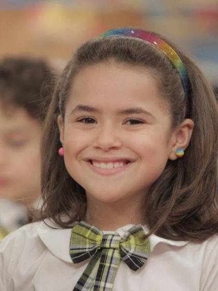 Garota sorridente com cabelo castanho, tiara colorida e uniforme escolar. Sorriso lembra alegria das novelas infantis.