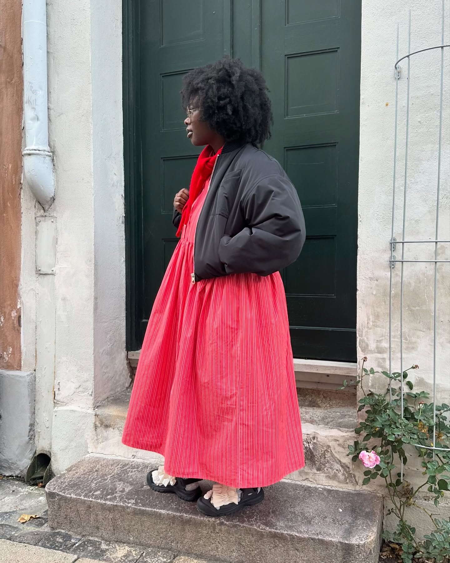 Pessoa usa vestido vermelho com casaco preto, posando em frente a uma porta verde.