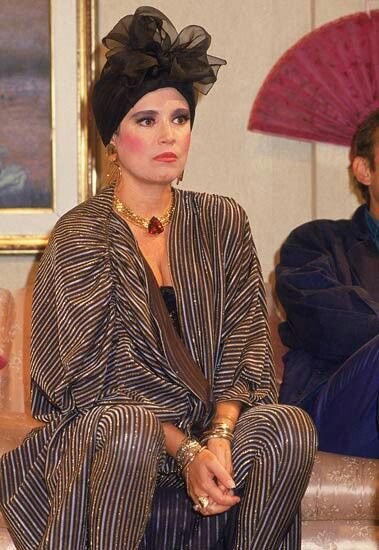 Mulher com roupa listrada, expressão pensativa e turbante preto, em cenário de novela.