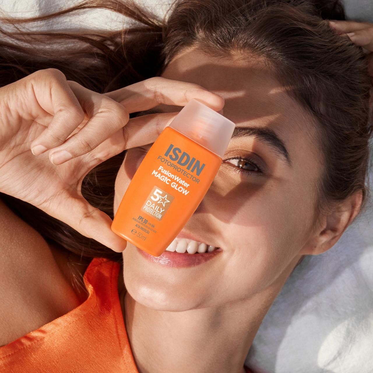 Mulher sorridente segura protetor solar Isdin Fusion Water Magic Glow na frente do rosto.