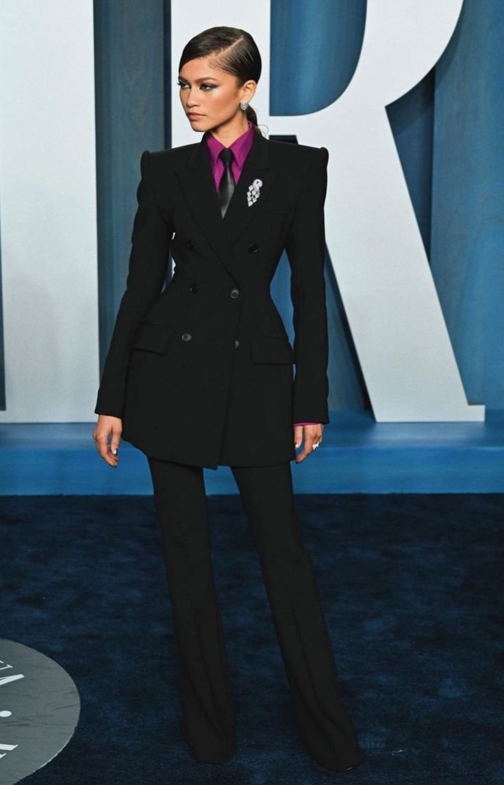 Pessoa usando terninho preto, camisa roxa e gravata preta em um evento de gala. Fundo azul com grande letra atrás.