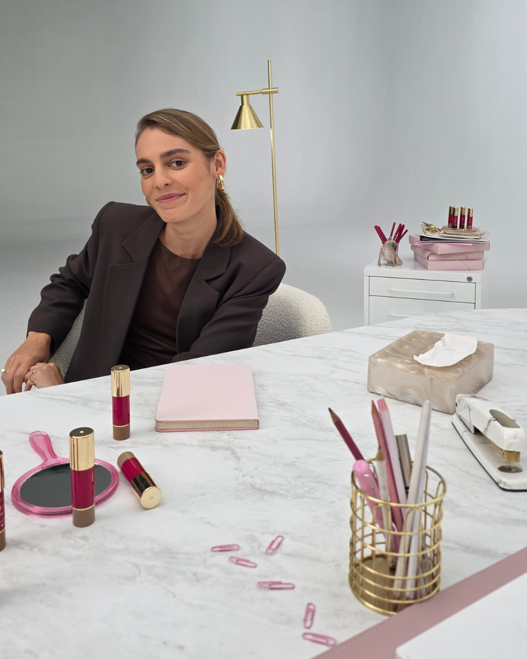 Mulher sentada em frente a uma mesa com produtos em stick, espelho e itens de escritório.