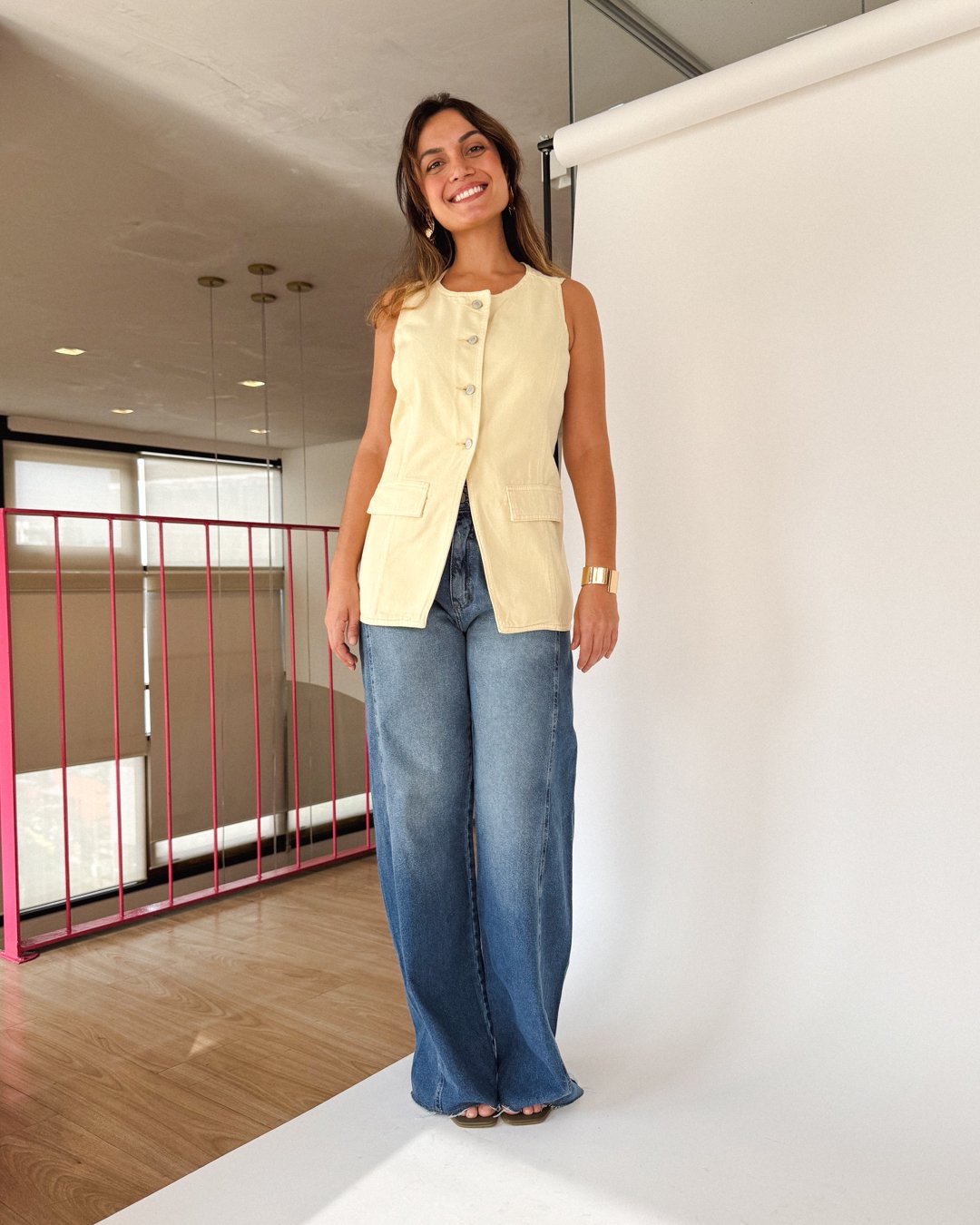 Mulher sorridente em colete bege e calça jeans, feitas em tecido mais sustentável em ambiente interno.