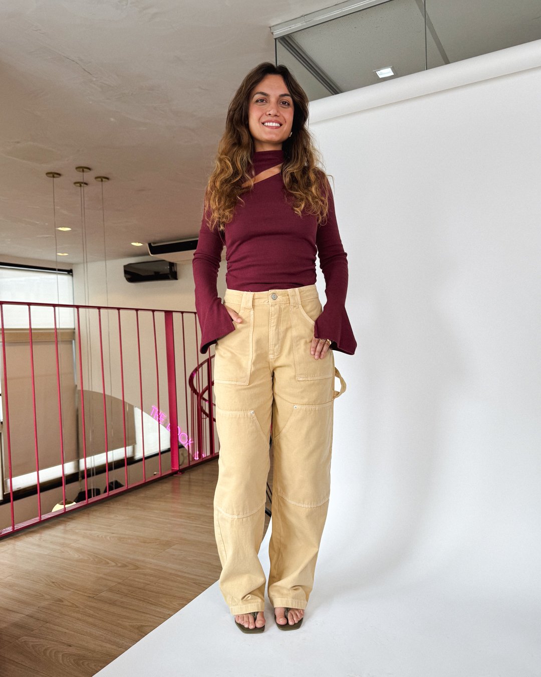Pessoa sorrindo, usando blusa vinho e calça bege, em um ambiente com piso de madeira e grade rosa ao fundo. Roupa feita de tecido mais sustentável