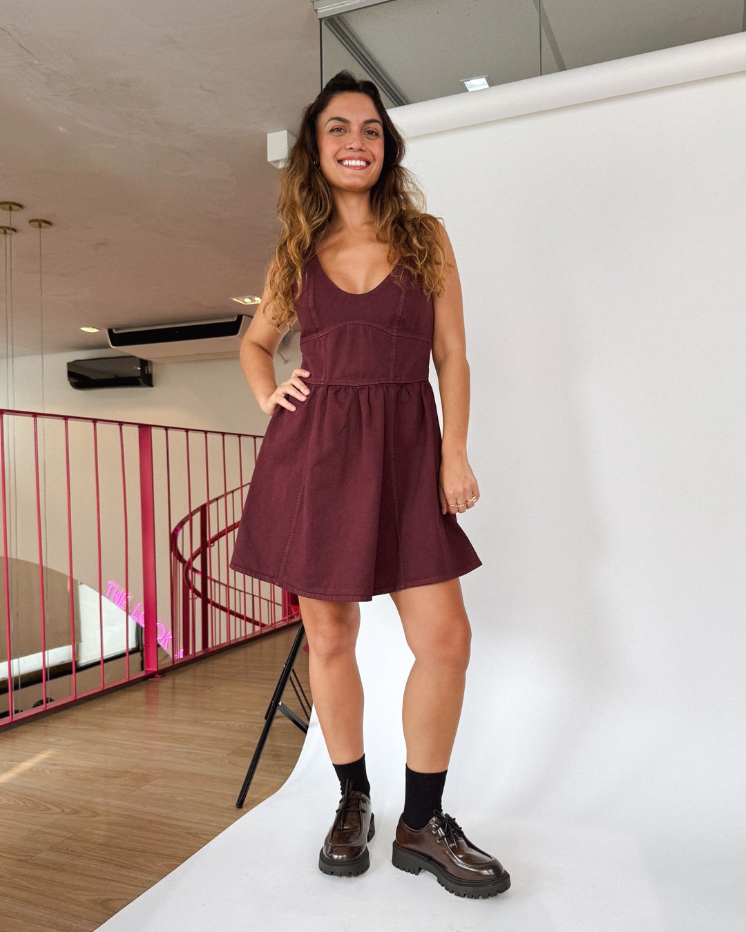 Modelo com vestido vinho, feito em tecido mais sustentável, sorriso, em estúdio com chão de madeira e estrutura metálica.