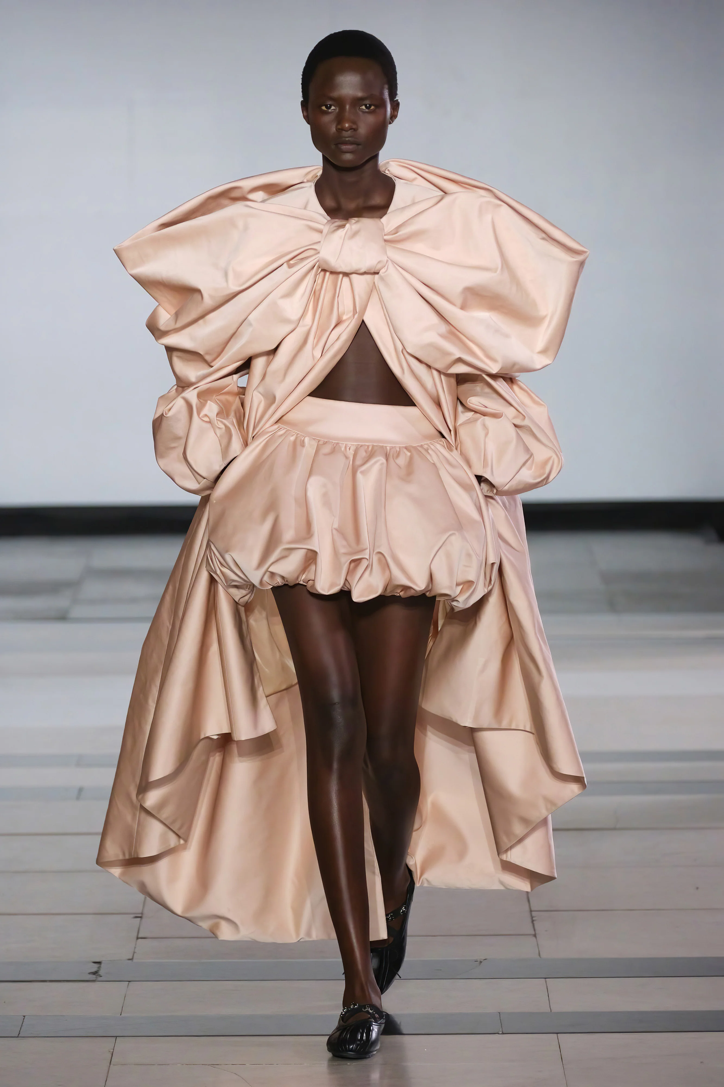 Modelo em desfile com vestido volumoso em tom nude, destacando tendência de beleza contemporânea.