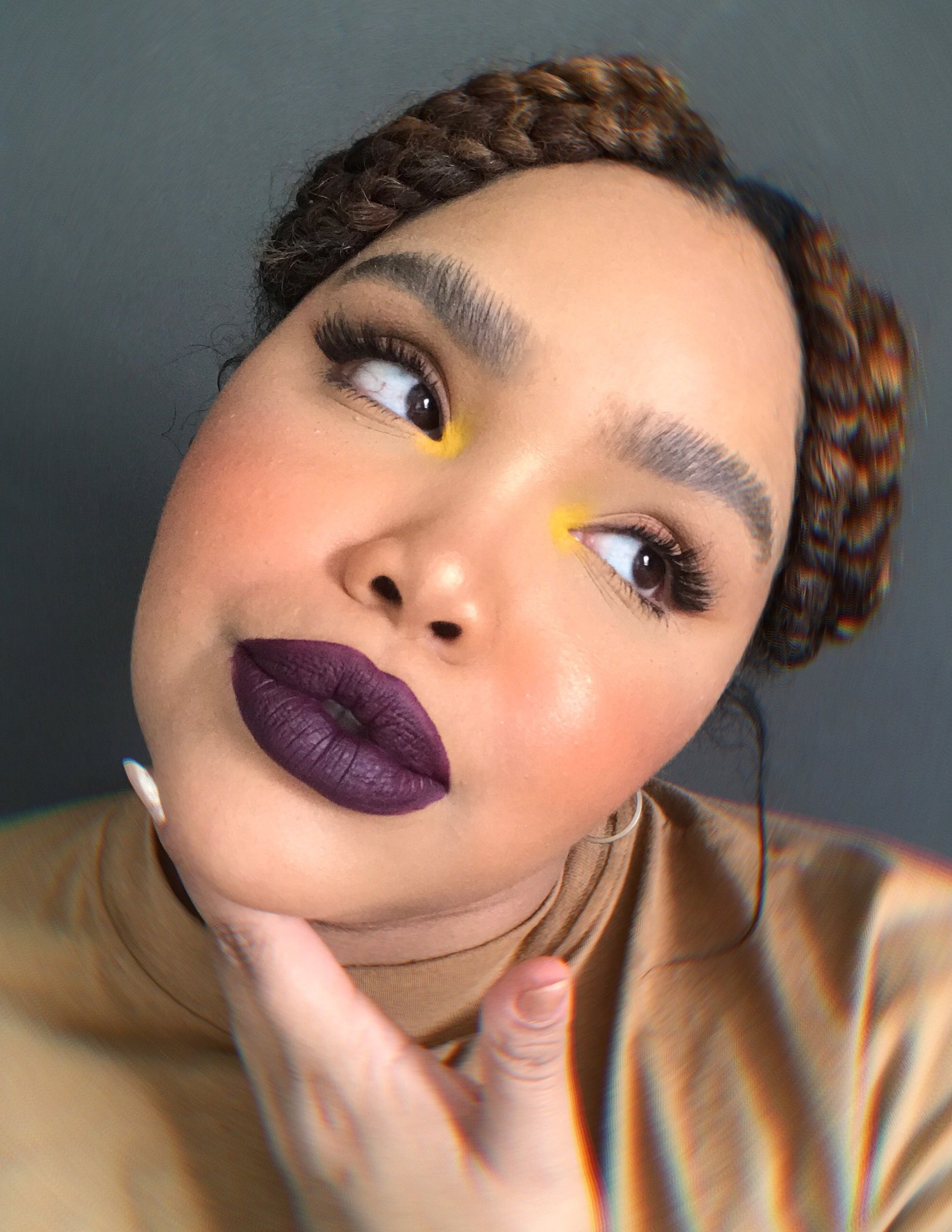 Mulher com maquiagem marcante, batom roxo e sombra amarela, posa de perfil com expressão pensativa.