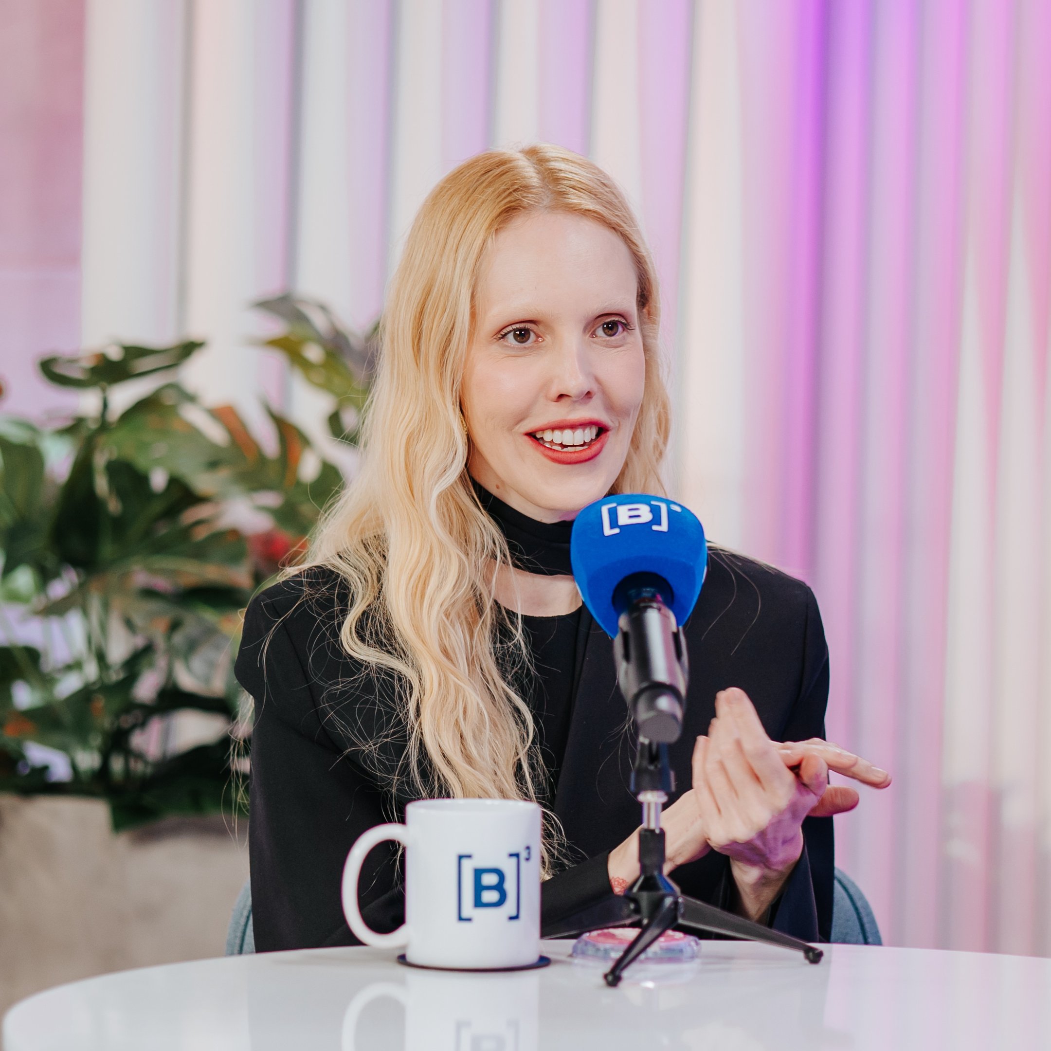 Jana Rosa fala ao microfone em estúdio decorado, destacando-se uma caneca sobre a mesa, contando sobre suas dívidas.