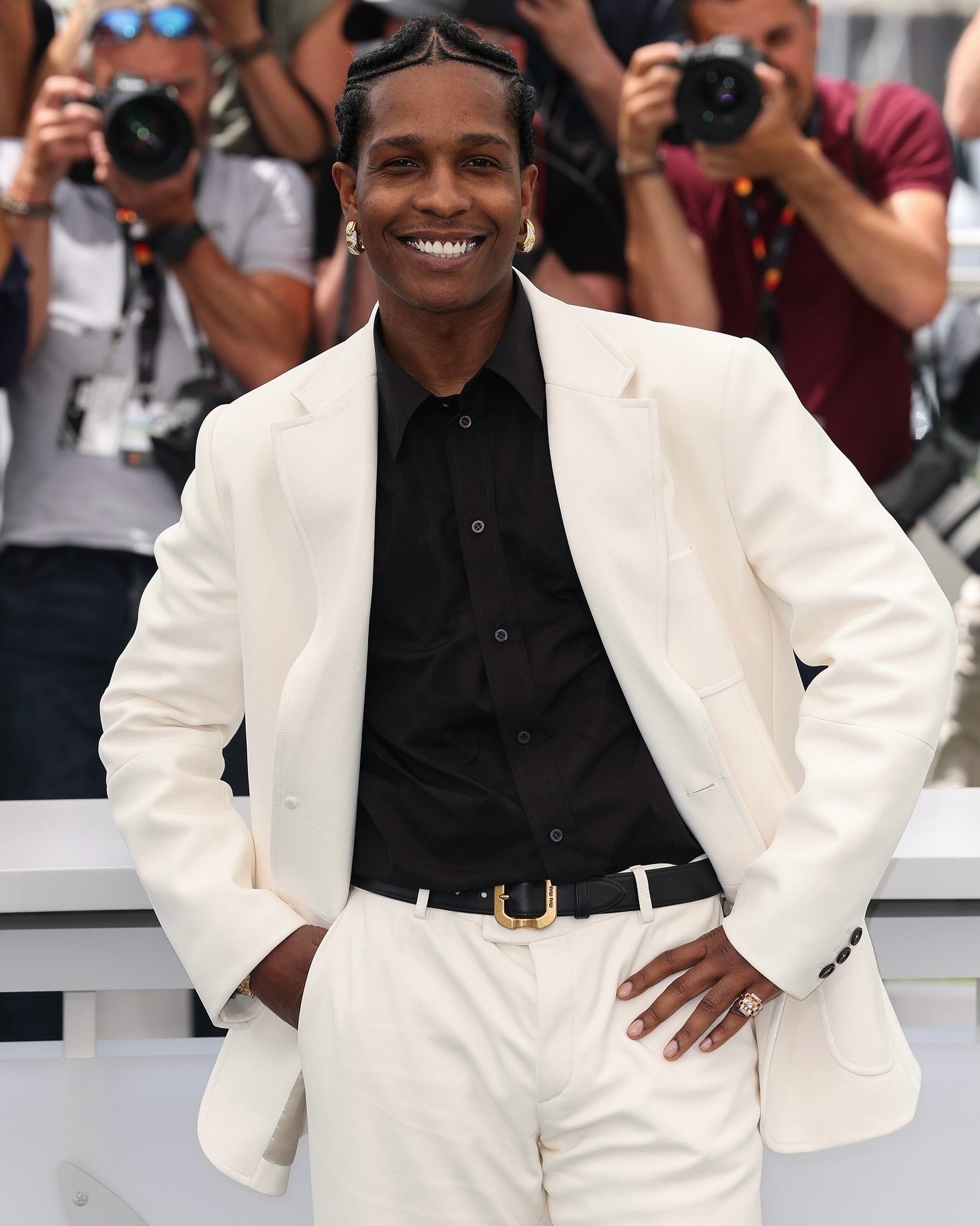 Sorridente em traje branco elegante no Festival de Cannes 2025, usando acessórios de destaque com fotógrafos ao fundo.