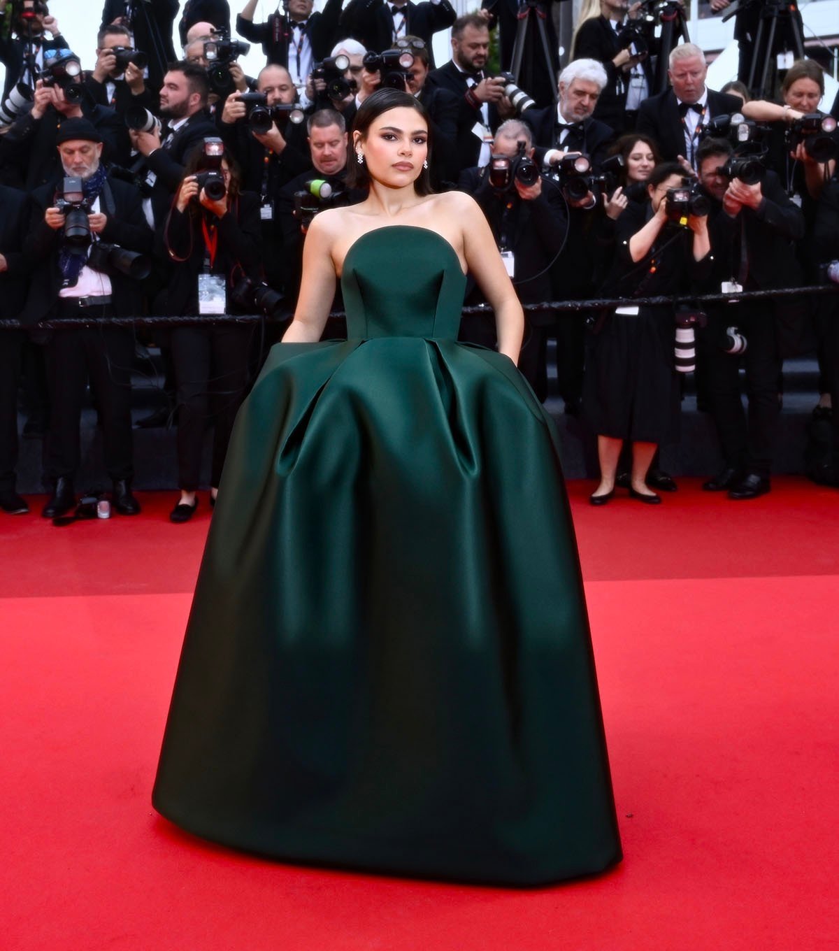 A pessoa está em um tapete vermelho, vestindo um longo vestido verde elegante, cercada por fotógrafos.