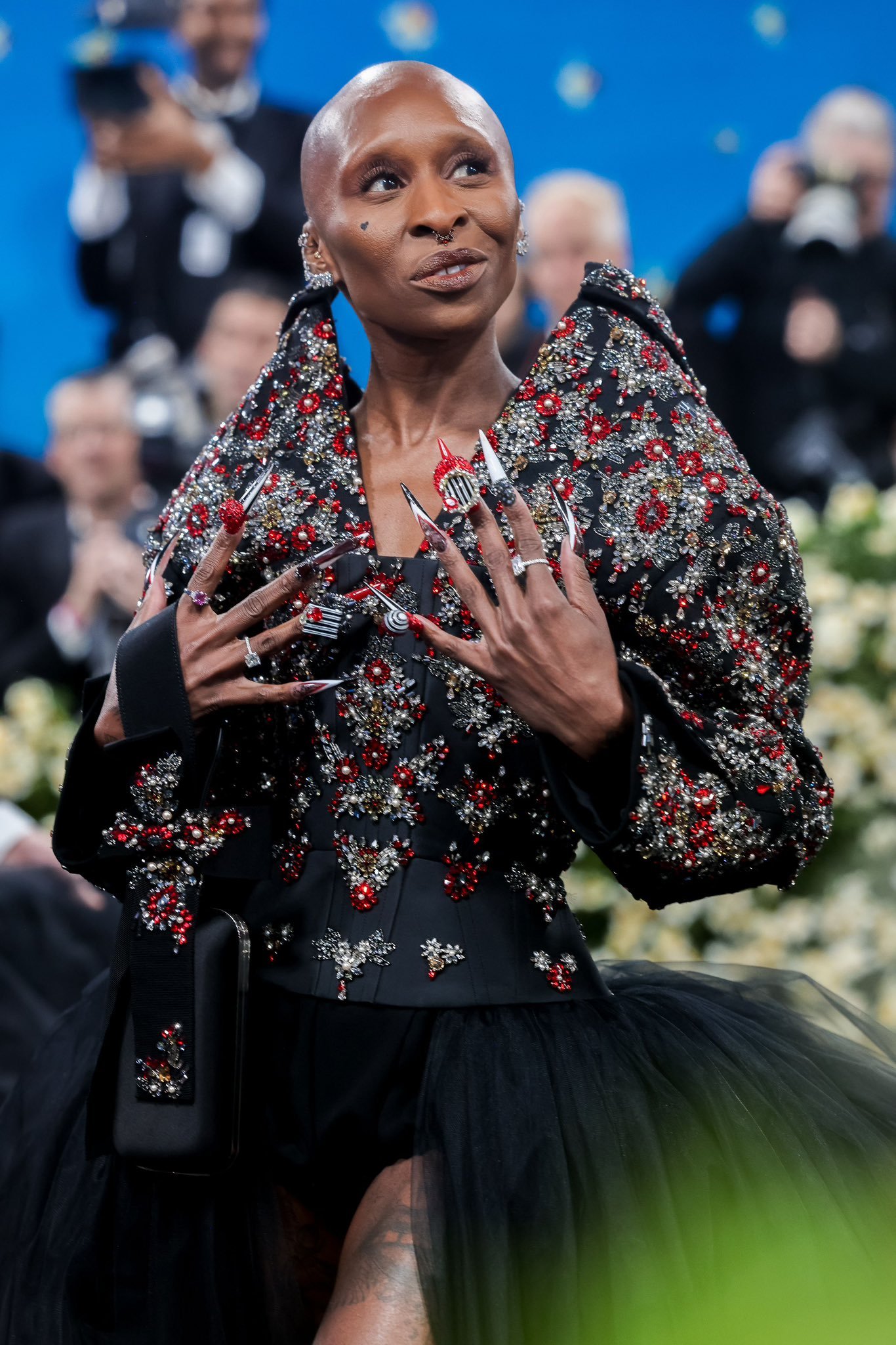 Celebridade em traje preto detalhado com pedras vermelhas e prateadas, mostrando design único e beleza icônica. Beleza das celebridades