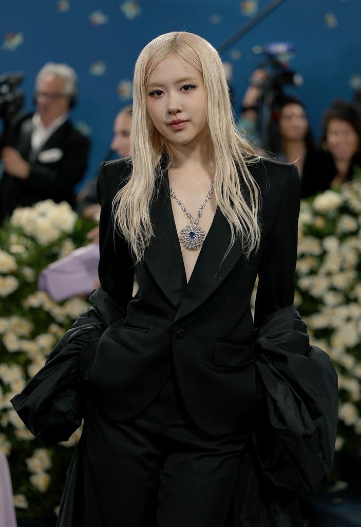 Celebridade em traje elegante e colar, cercada por flores e pessoas ao fundo, realçando a beleza das celebridades.