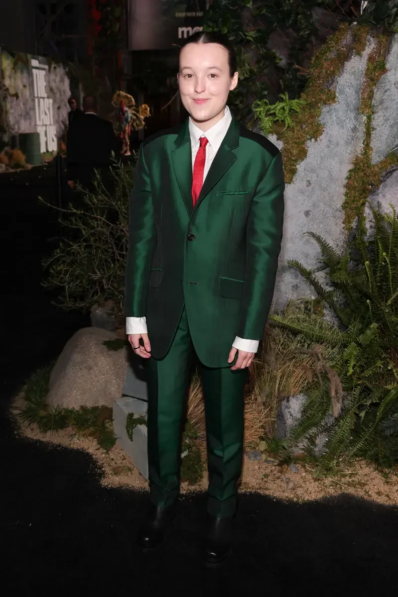 Bella Ramsey em traje verde escuro, com gravata vermelha, em pé perto de folhagens e rochas decorativas.