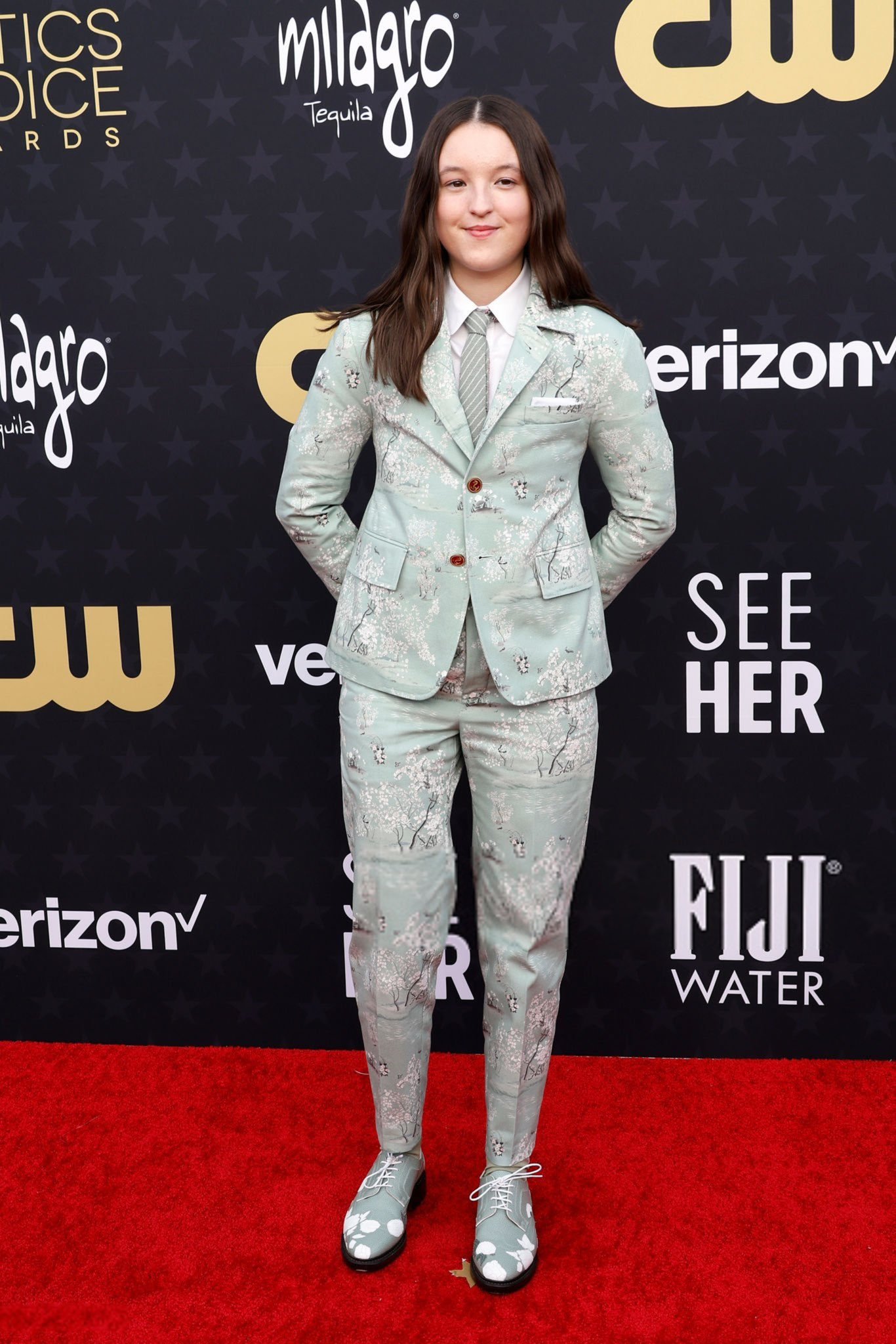 Bella Ramsey está em um tapete vermelho, usando um terno claro com estampa floral.