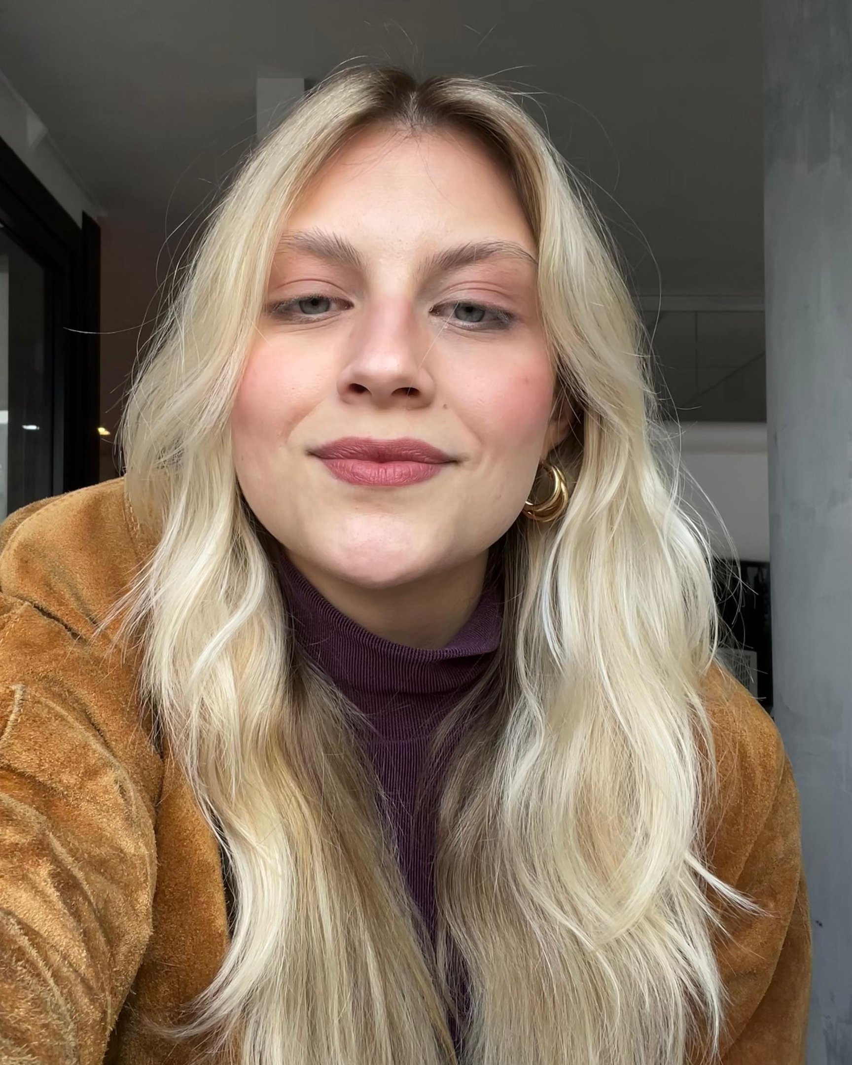 Pessoa com cabelo loiro e maquiagem discreta, incluindo blush Vizzela. Ela usa casaco marrom e suéter roxo.