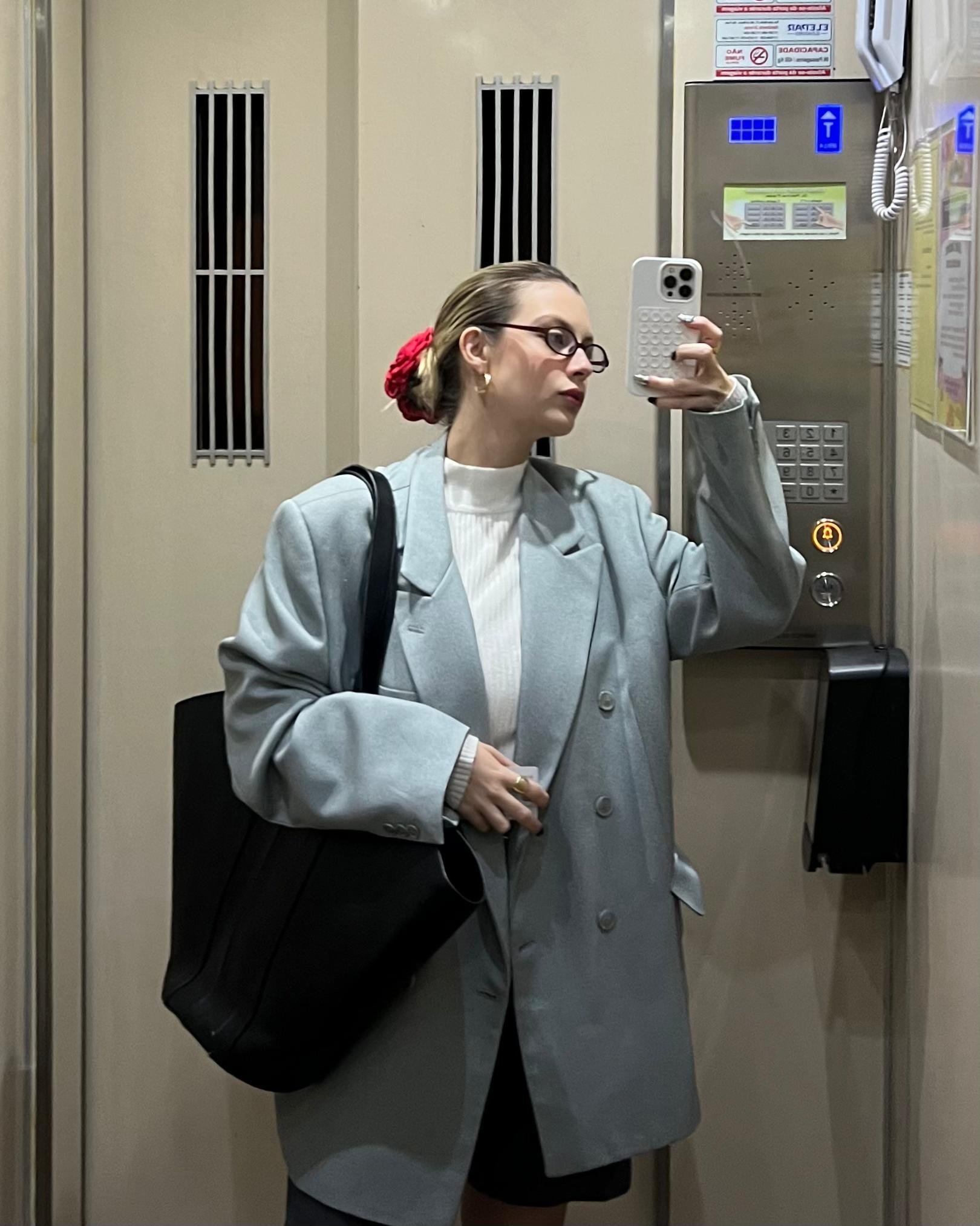 Mulher em elevador usa blazer cinza, bolsa de adulto preta no ombro e tira selfie no espelho.