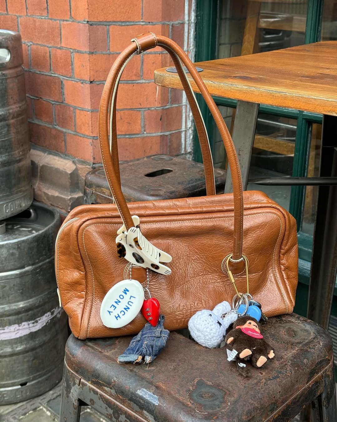 Bolsa feminina em couro marrom com chaveiros variados pendurados, apoiada em uma superfície metálica.